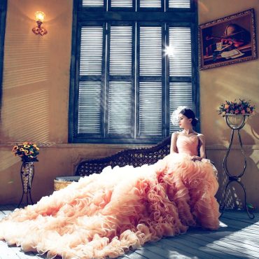A bride in a luxurious gown sits gracefully in a sunlit room, exuding elegance.