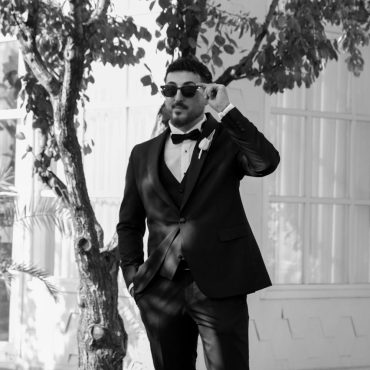 A man in a tuxedo adjusts his sunglasses outdoors, exuding elegance and style.